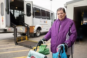 Photo of a person using a walker standing in front of a bus with a wheelchair lift.