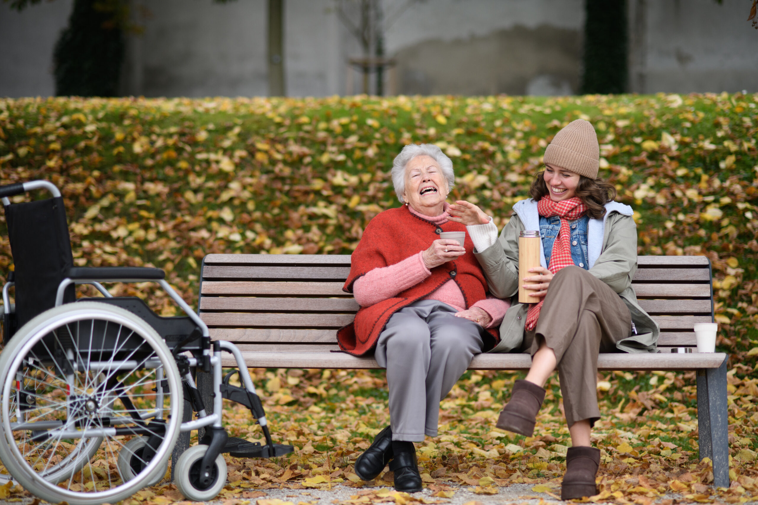 Smiling woman caregiver sharing a beverage with laughing senior woman on park bench.