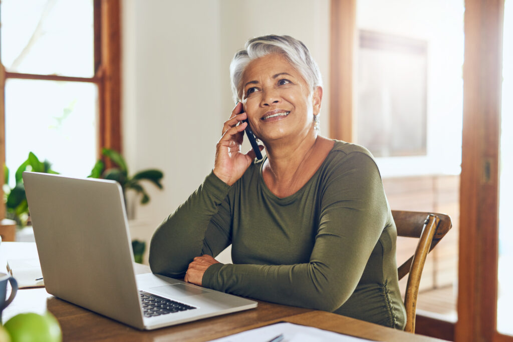Senior woman of Asian descent sitting with laptop and talking on cellphone.