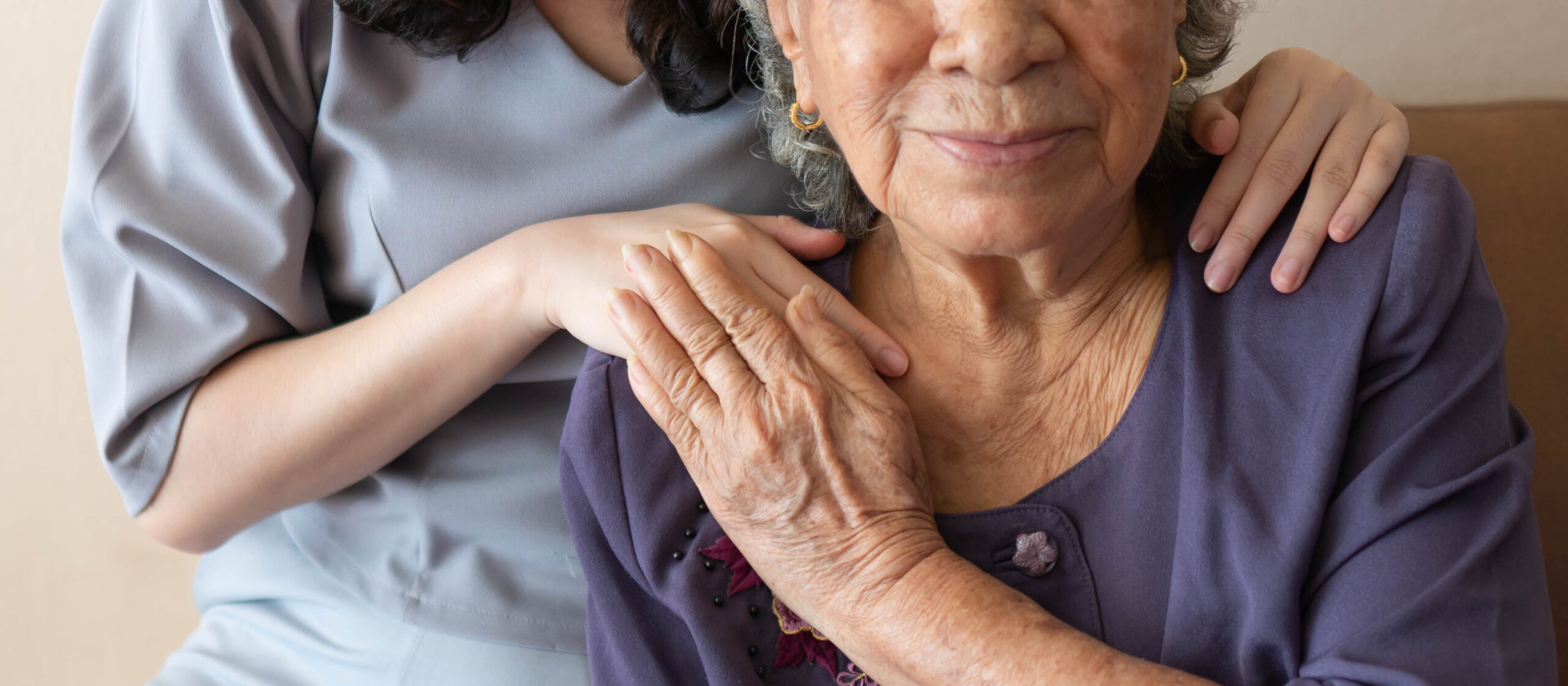 Photo of an older woman sitting down. A younger woman stands behind her. They are holding hands.