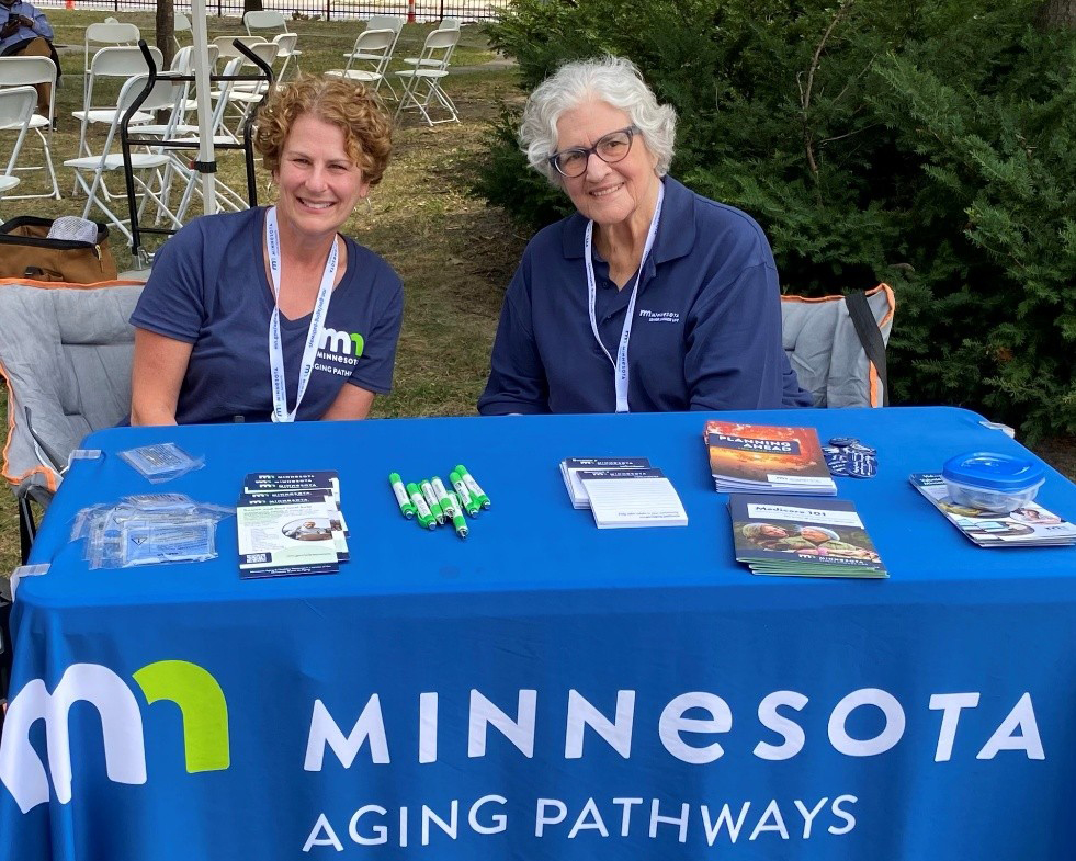 Two volunteers sitting at a Minnesota Aging Pathways information table at an outdoor event.